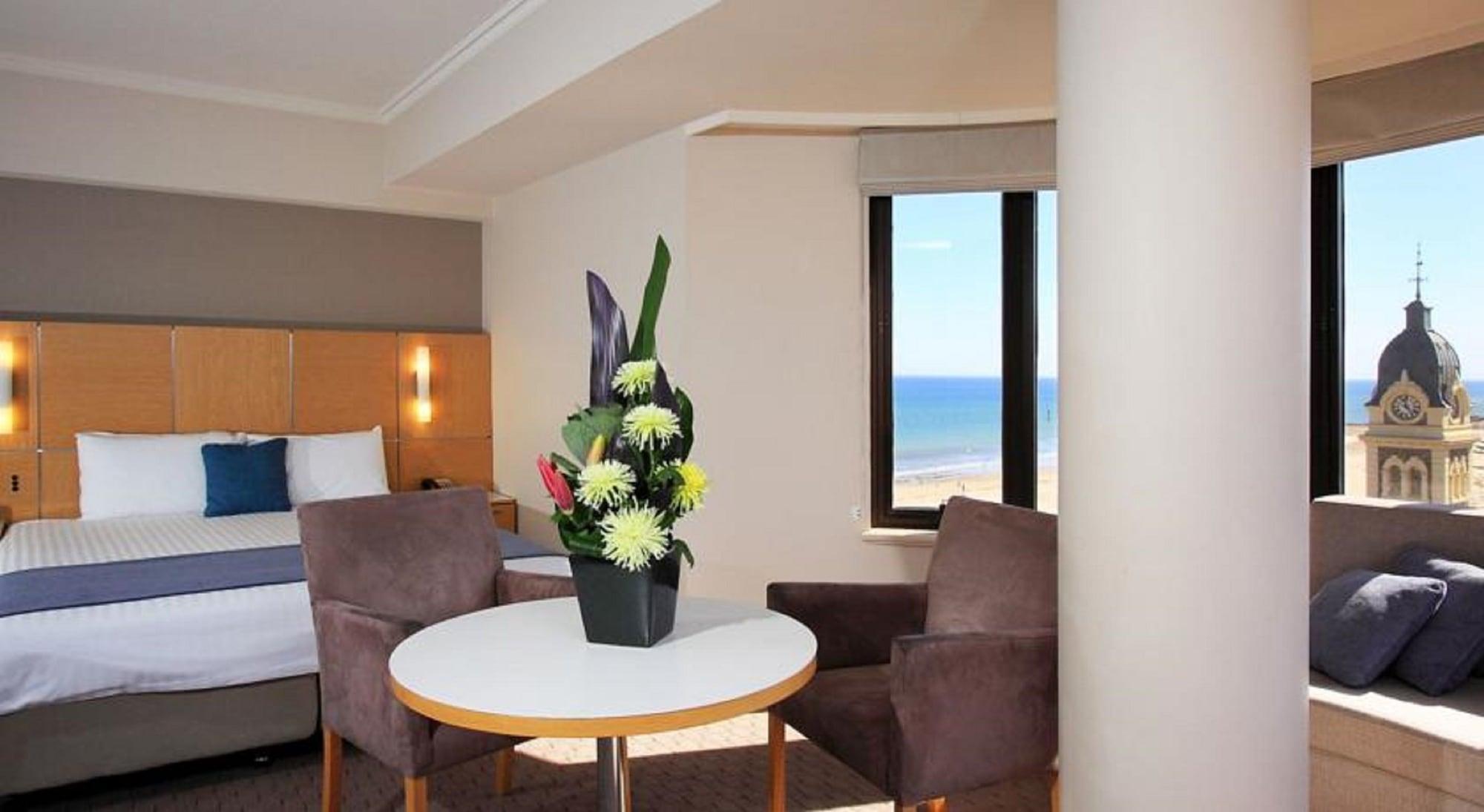 Stamford Grand Adelaide Otel Glenelg Dış mekan fotoğraf The photo shows a cozy hotel room with a comfortable seating arrangement. There are two brown chairs positioned around a round table, which features a decorative flower arrangement in a black vase. The background includes a window that offers a view 