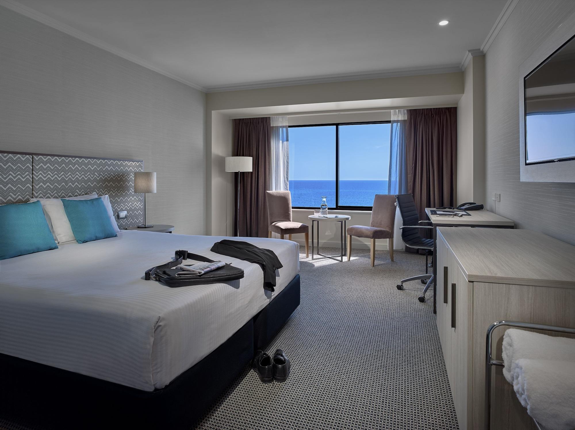 Stamford Grand Adelaide Otel Glenelg Dış mekan fotoğraf The photo shows a modern hotel room featuring a queen-sized bed with white linens and two aqua blue decorative pillows. There are bedside lamps and a large window with a view of the ocean. The room also has a small seating area with two chairs and a 