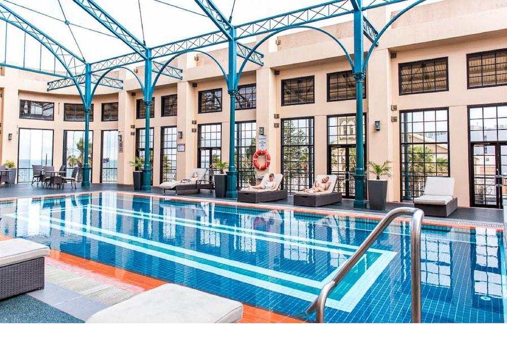 Stamford Grand Adelaide Otel Glenelg Dış mekan fotoğraf The photo shows an indoor swimming pool area characterized by a modern and elegant design. The pool features blue tiles with two white stripes running along its length. Surrounding the pool, there are several lounge chairs for relaxation. The walls h