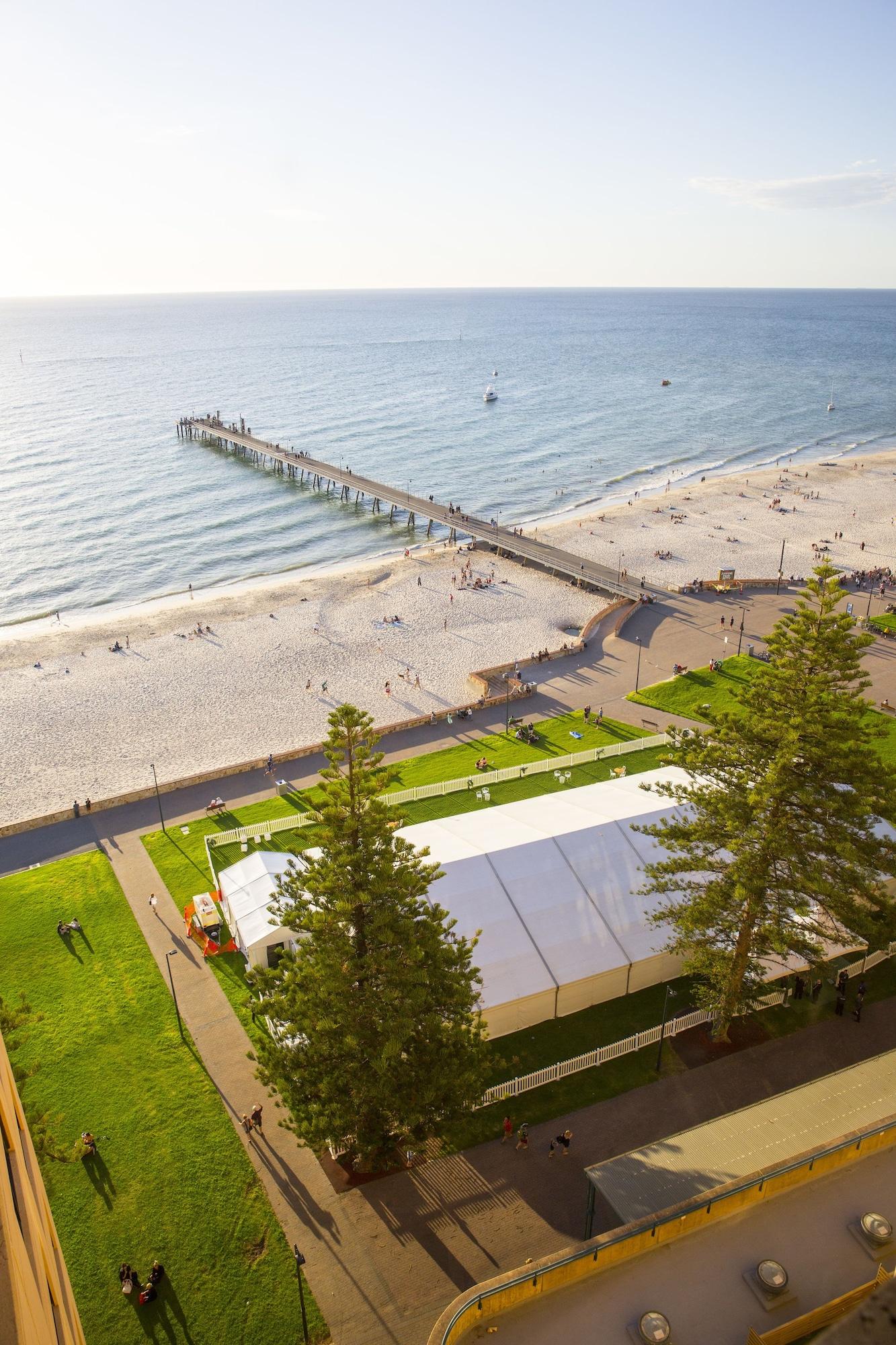 Stamford Grand Adelaide Otel Glenelg Dış mekan fotoğraf The photo shows a scenic view of a beach with a long pier extending into the water. The beach is sandy, and there are people enjoying the sun and walking along the shore. In the foreground, there is a large white tent set up on a grassy area, likely 