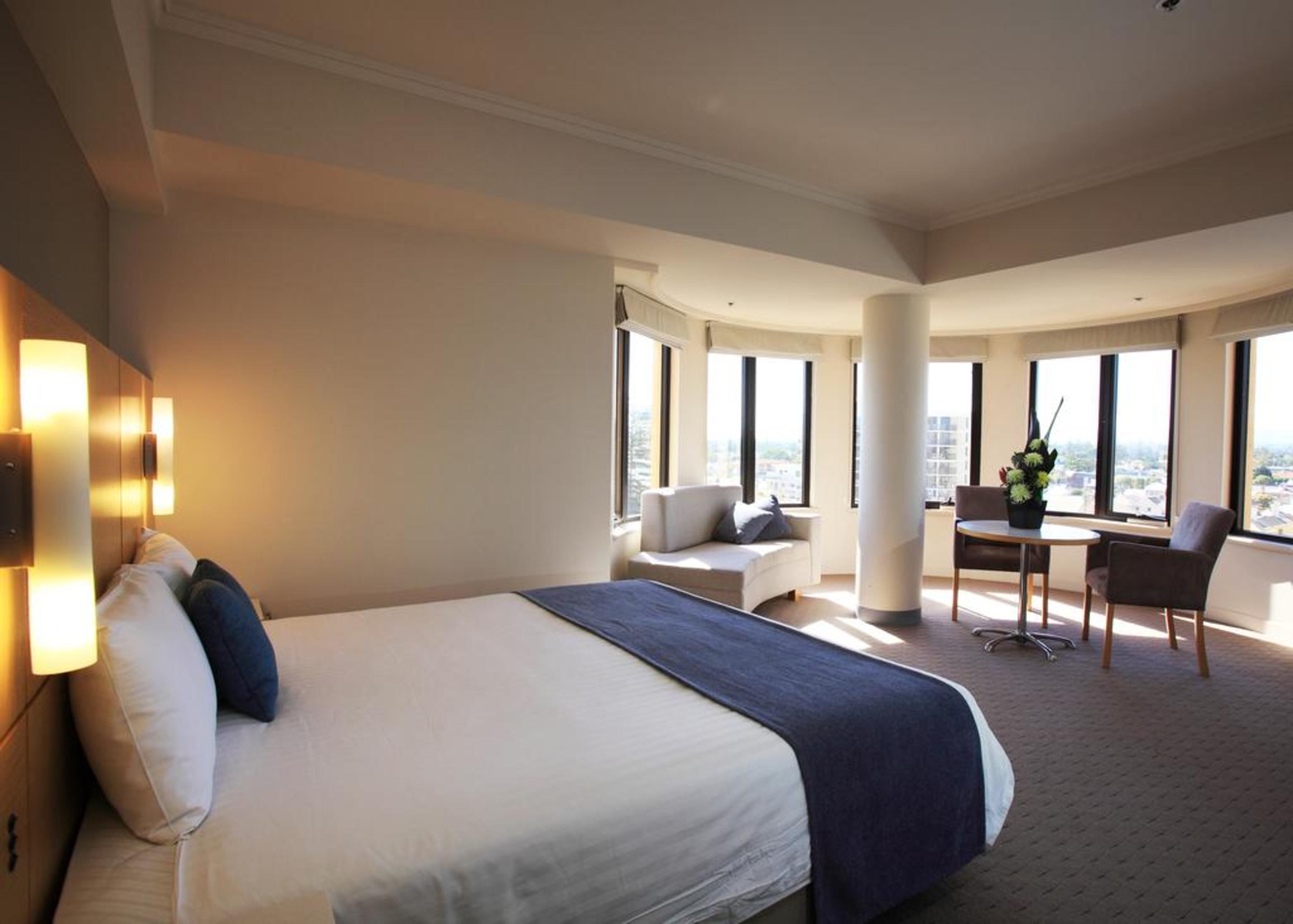 Stamford Grand Adelaide Otel Glenelg Dış mekan fotoğraf The photo shows a hotel room with a modern and spacious design. There is a large bed with white linens and a dark blue throw blanket. The room features multiple windows that allow plenty of natural light, and there is a small seating area near the wi