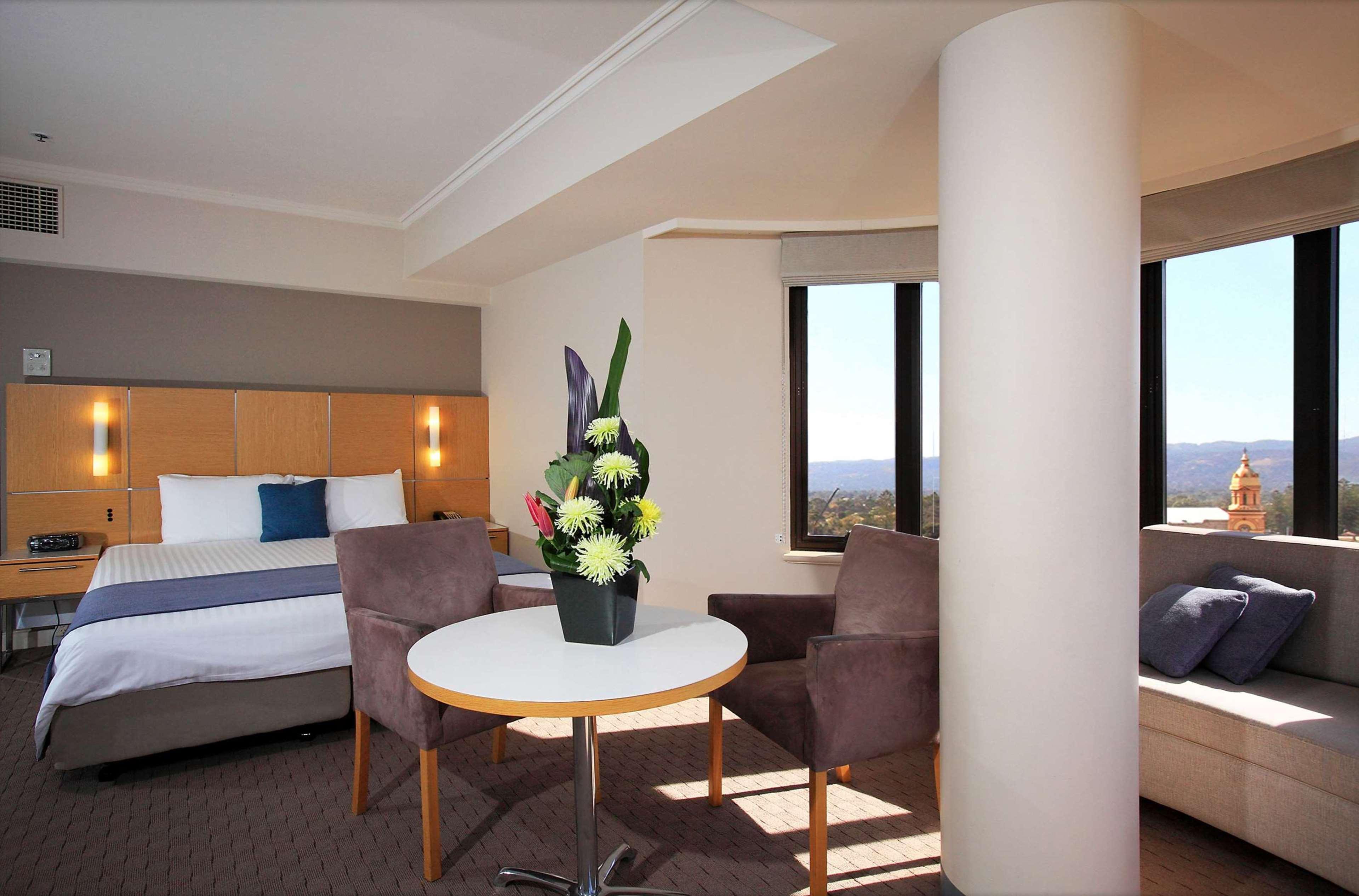Stamford Grand Adelaide Otel Glenelg Dış mekan fotoğraf The photo shows a hotel room with a modern design. On one side, there is a plush bed with white and blue bedding. In the center of the room, there's a small round table with two gray upholstered chairs around it. A vase of flowers sits on the table, 