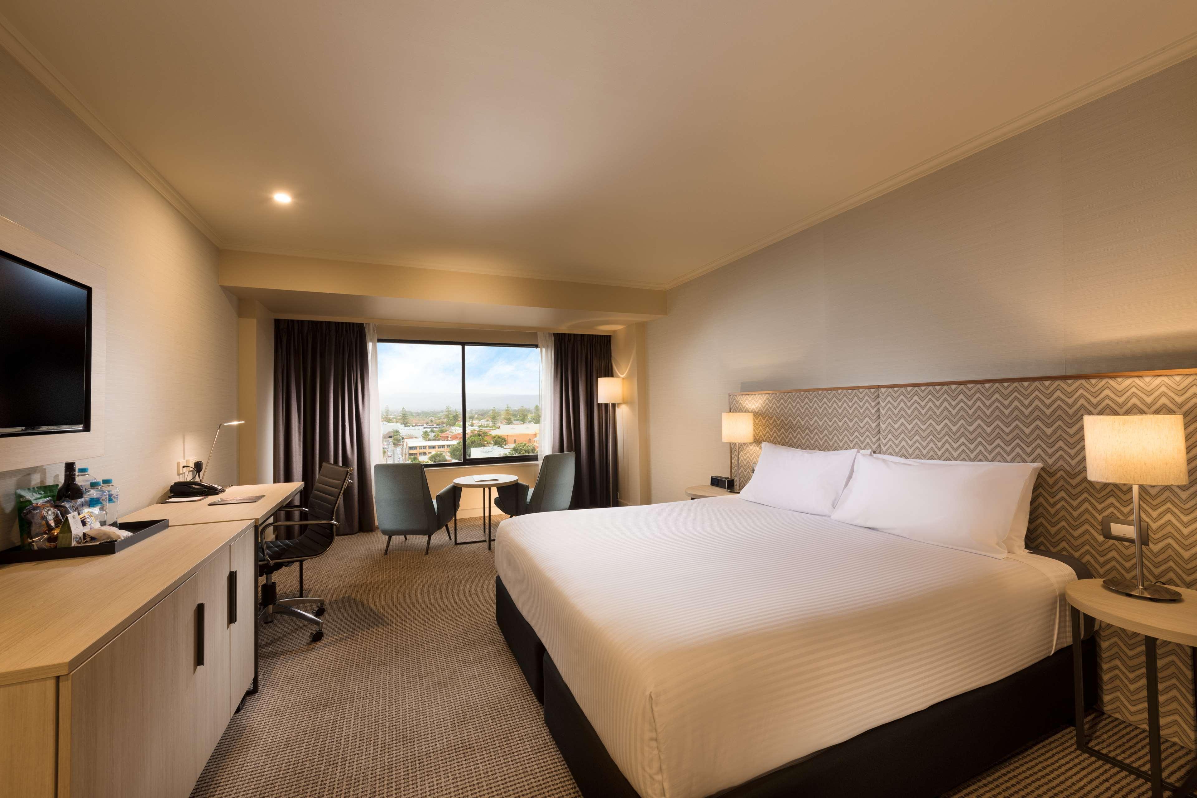 Stamford Grand Adelaide Otel Glenelg Dış mekan fotoğraf The photo shows a modern hotel room. It features a large bed with white linens, a desk with a chair, and a flat-screen TV mounted on the wall. There are two armchairs and a small table near a large window that lets in natural light, providing a view 