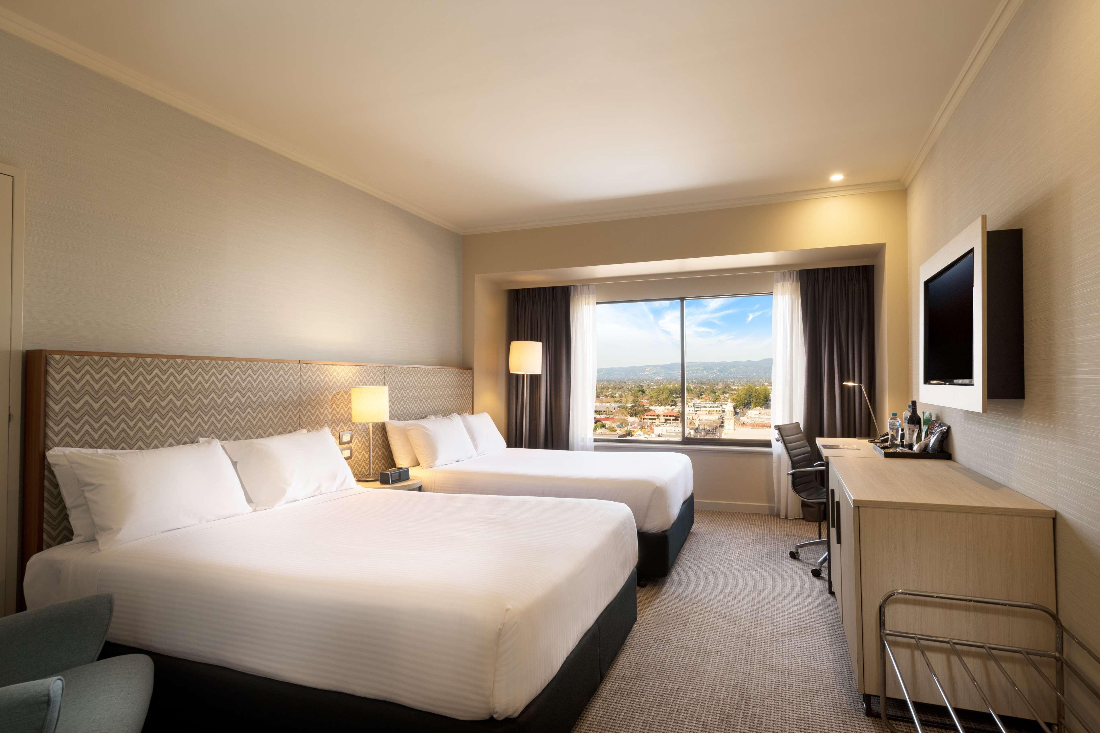 Stamford Grand Adelaide Otel Glenelg Dış mekan fotoğraf The photo shows a modern hotel room featuring two beds with white linens. There are bedside tables with lamps next to each bed. A large window provides natural light and offers a view of the outside, with curtains on either side. The room has a neutr