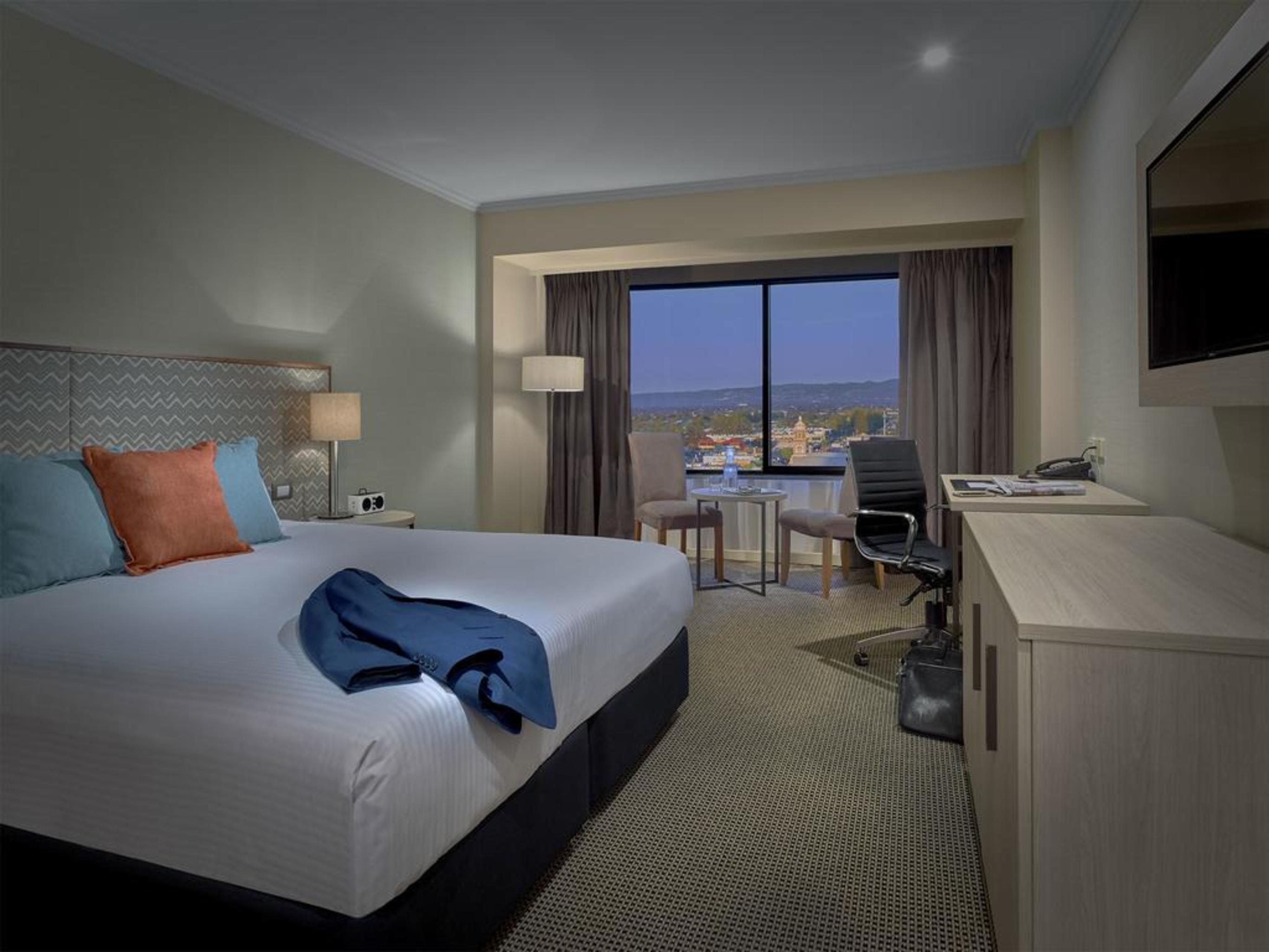 Stamford Grand Adelaide Otel Glenelg Dış mekan fotoğraf The photo shows a modern hotel room. It features a large double bed with white bedding and decorative pillows in orange and blue. There's a nightstand with lamps on either side of the bed. A large window overlooks a view outside, and there's a desk a