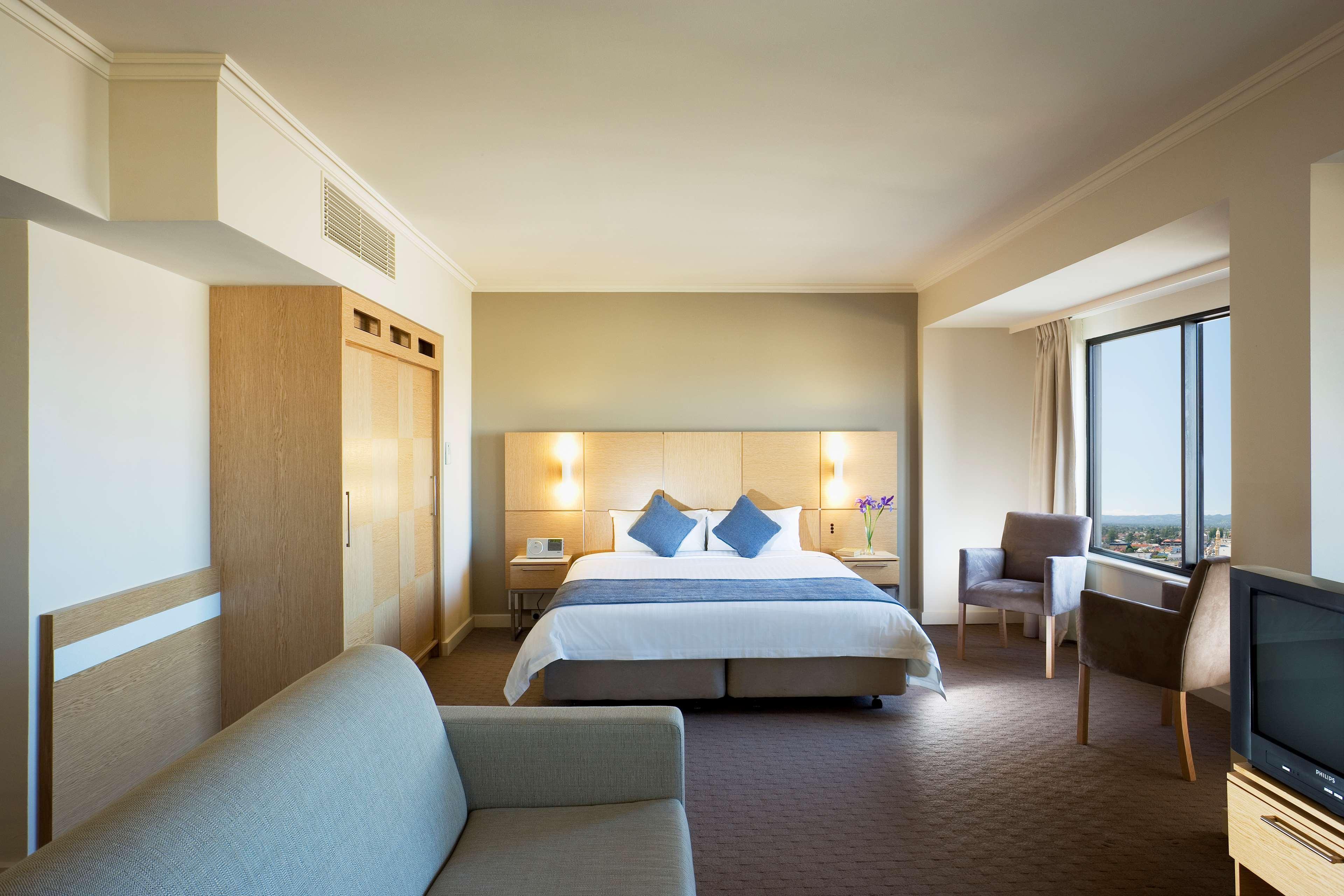 Stamford Grand Adelaide Otel Glenelg Oda fotoğraf The photo shows a modern hotel room featuring a large bed with two blue decorative pillows. There are bedside lamps on both sides of the headboard. To the left, there's a wooden wardrobe and a small table with a vase and a few amenities. On the right