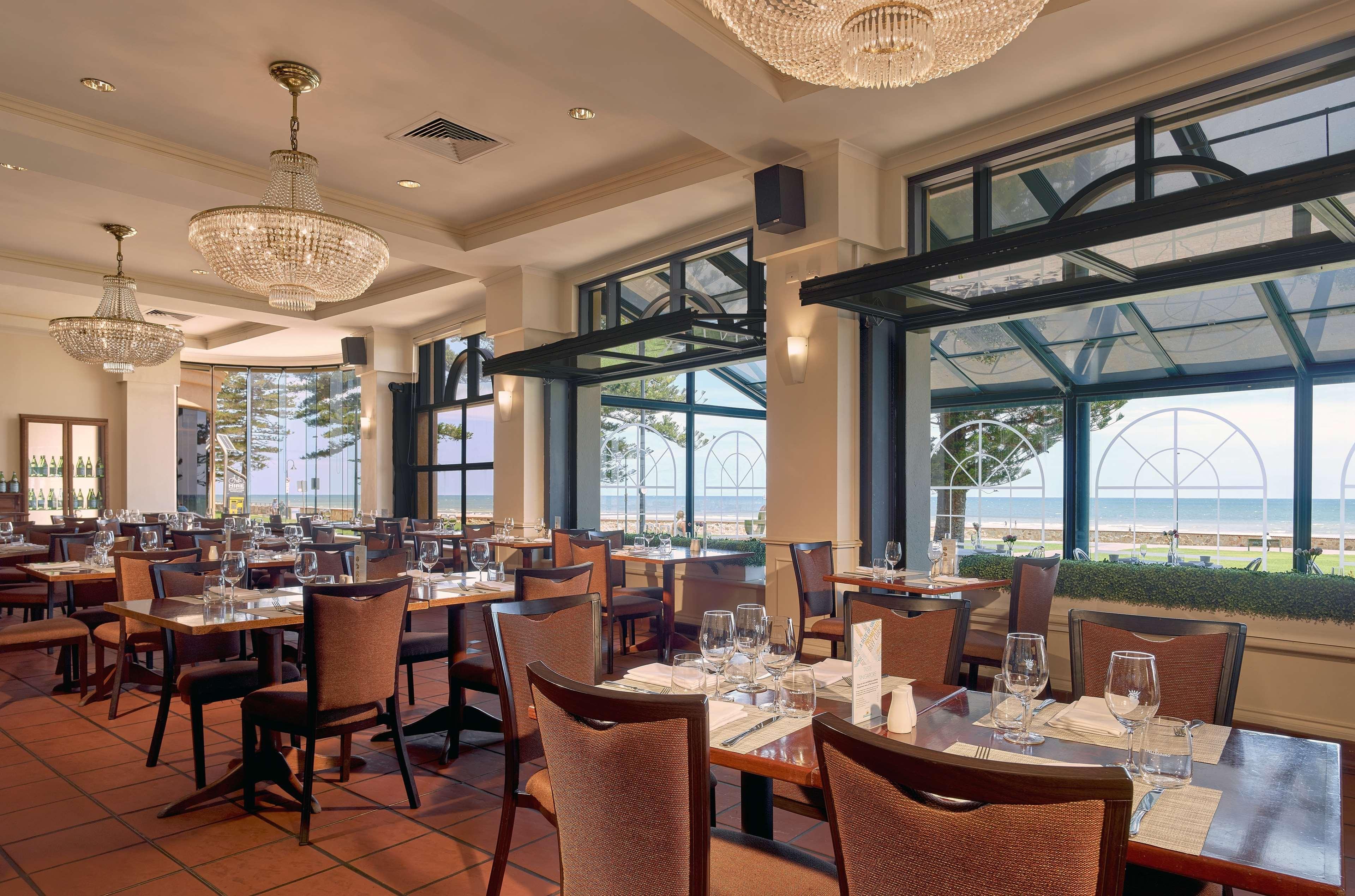Stamford Grand Adelaide Otel Glenelg Dış mekan fotoğraf The photo shows the interior of a restaurant. It features several wooden tables set with glasses and utensils, suggesting it's ready for diners. The decor includes elegant chandeliers hanging from the ceiling, adding a touch of sophistication. Large 