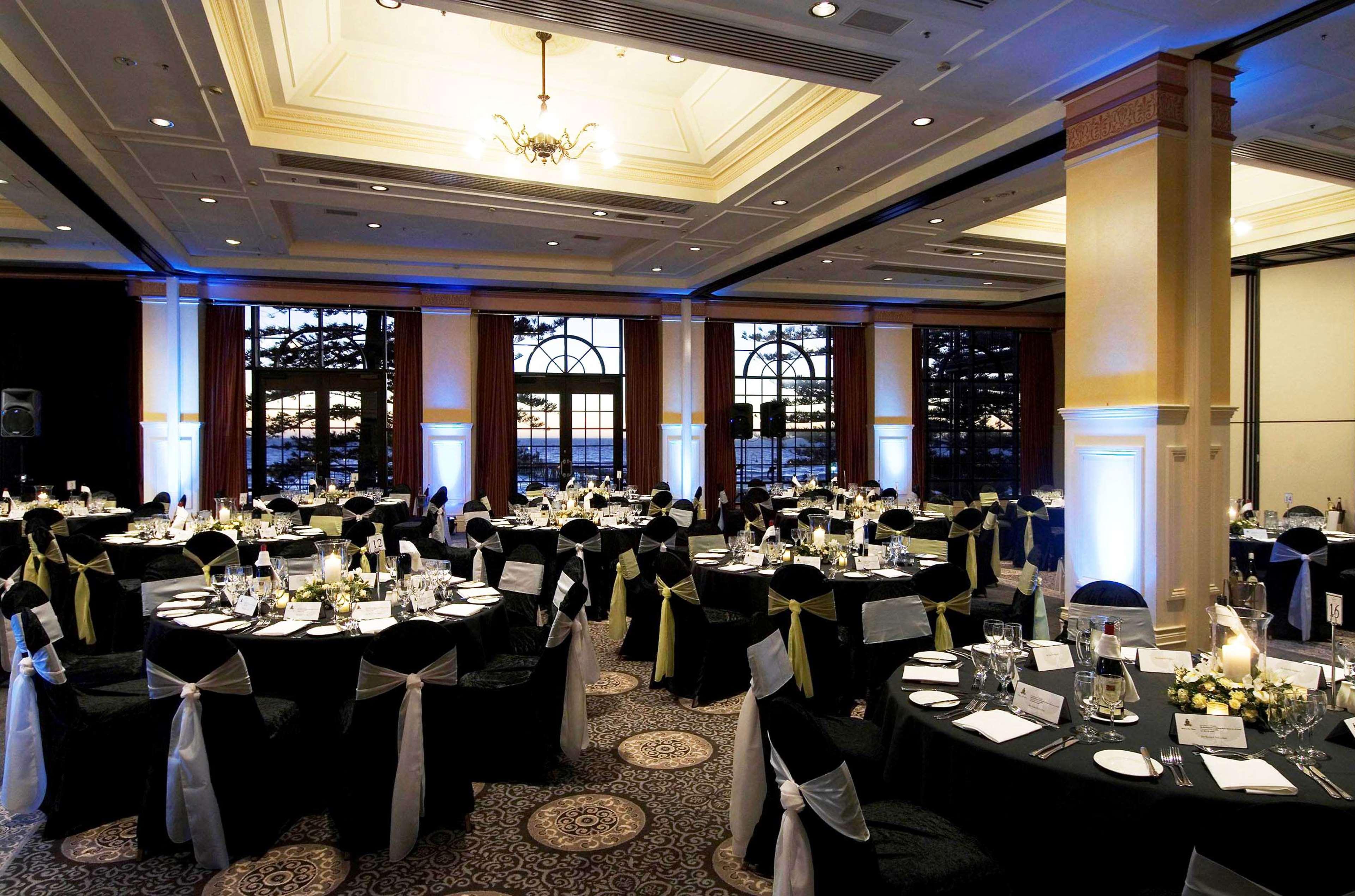 Stamford Grand Adelaide Otel Glenelg Restoran fotoğraf The photo depicts an elegantly arranged banquet hall set for an event. The room features round tables covered with black tablecloths, each adorned with white tableware and floral centerpieces. Chairs are draped in white fabric with decorative sashes 
