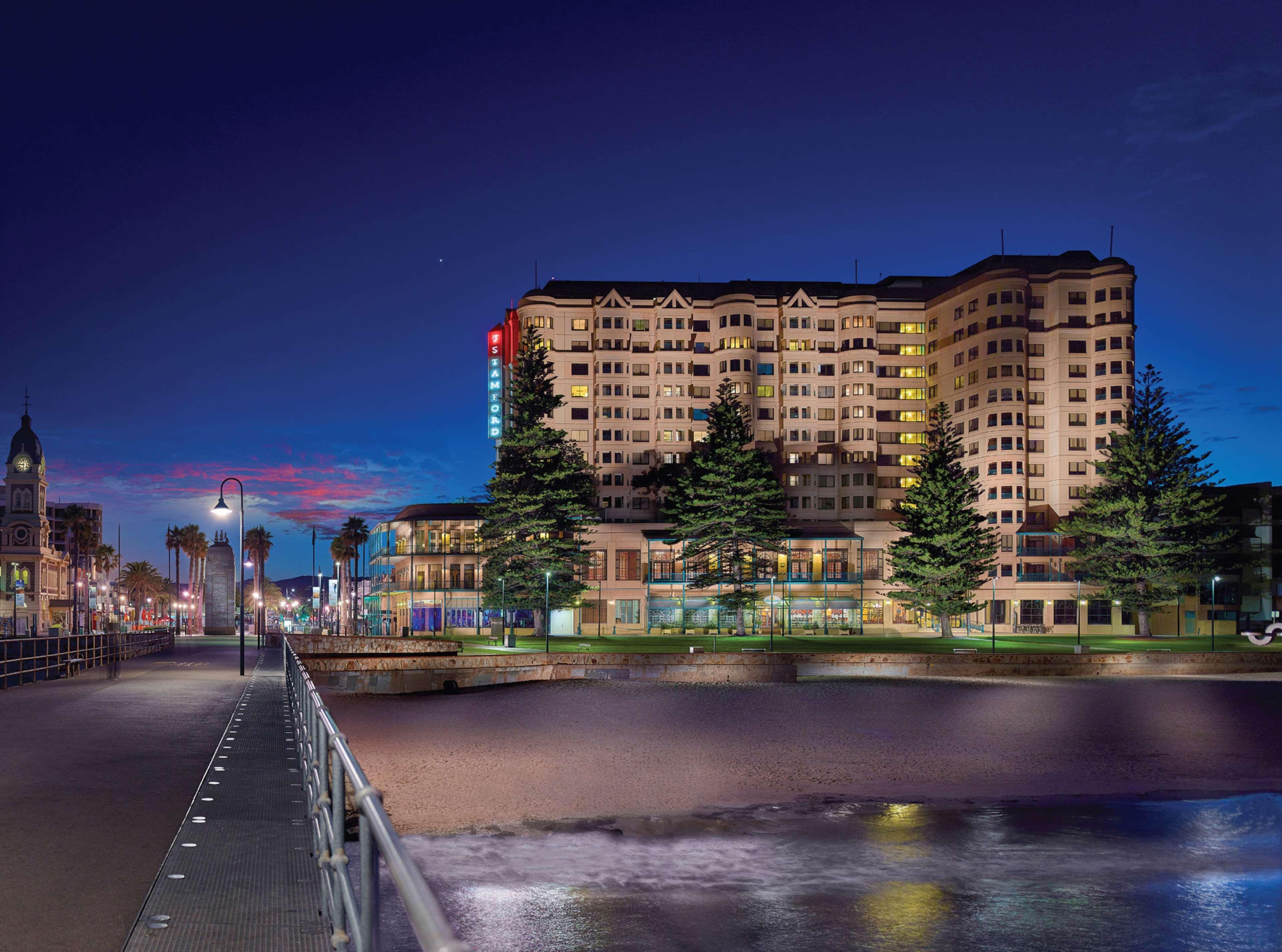 Stamford Grand Adelaide Otel Glenelg Dış mekan fotoğraf The photo shows a waterfront scene at dusk, featuring a large building lit up against a darkening sky. The architecture appears modern and prominent, with several windows illuminated. In the foreground, there is a walkway or pier with fencing along t
