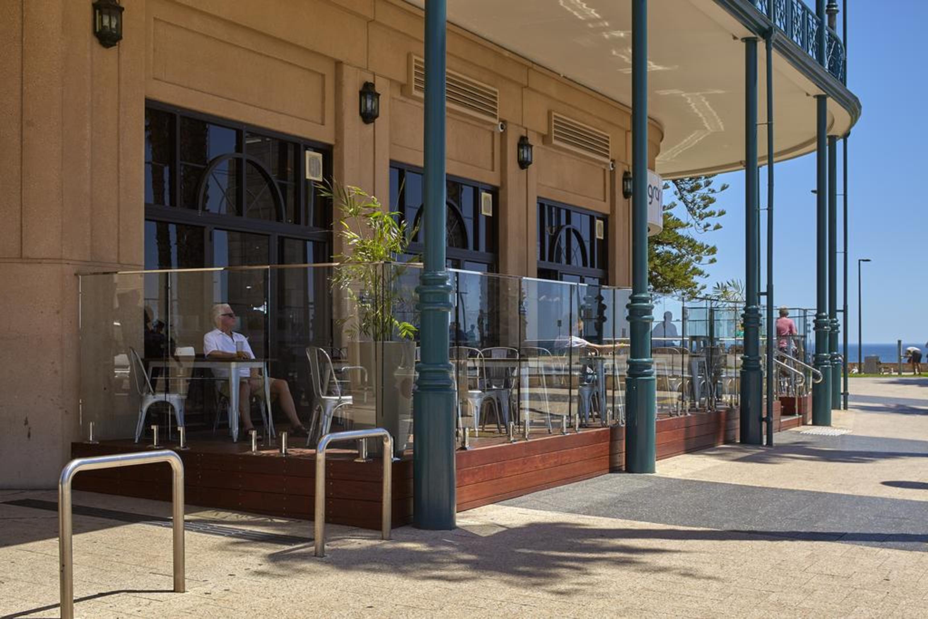 Stamford Grand Adelaide Otel Glenelg Dış mekan fotoğraf The photo shows the exterior of a cafe or restaurant with a large outdoor seating area. There are glass barriers around the seating, providing a view of the outside while maintaining a sense of enclosure. The building has a modern architectural style