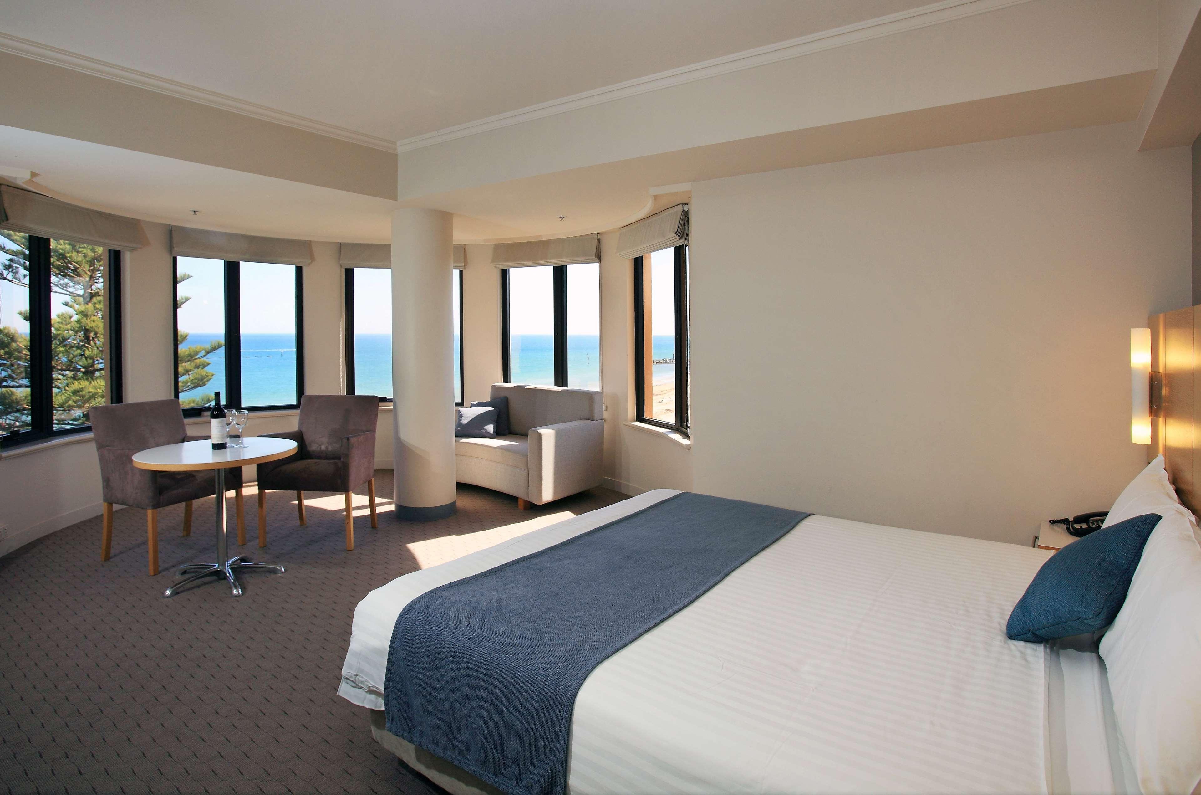 Stamford Grand Adelaide Otel Glenelg Dış mekan fotoğraf The photo shows a well-lit hotel room featuring large windows that offer a view of the ocean. The room is arranged with a comfortable bed covered in white linens and a navy blue throw blanket. There is a small table and chairs set up beside the windo
