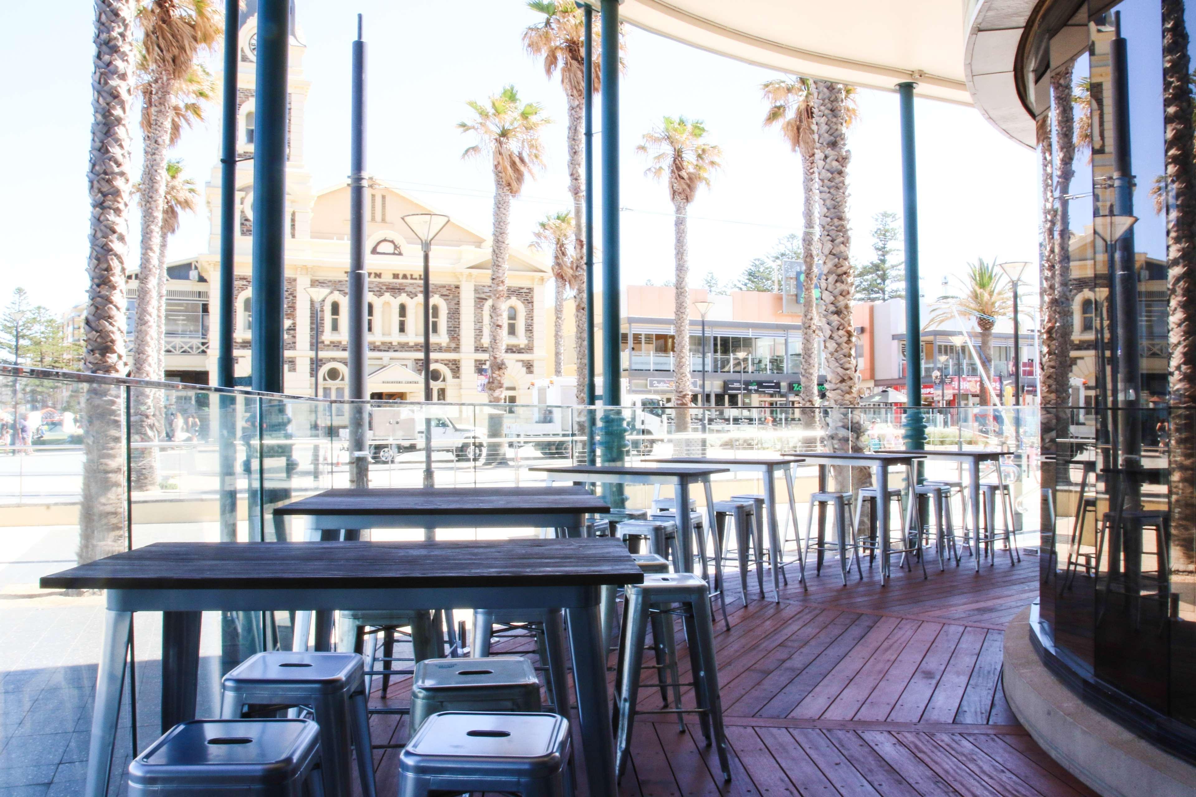 Stamford Grand Adelaide Otel Glenelg Dış mekan fotoğraf The photo shows an outdoor seating area of a café or restaurant. It features metal tables and stools arranged on a wooden deck. The setting has large glass panels, providing an open and airy atmosphere. In the background, palm trees and a building wi