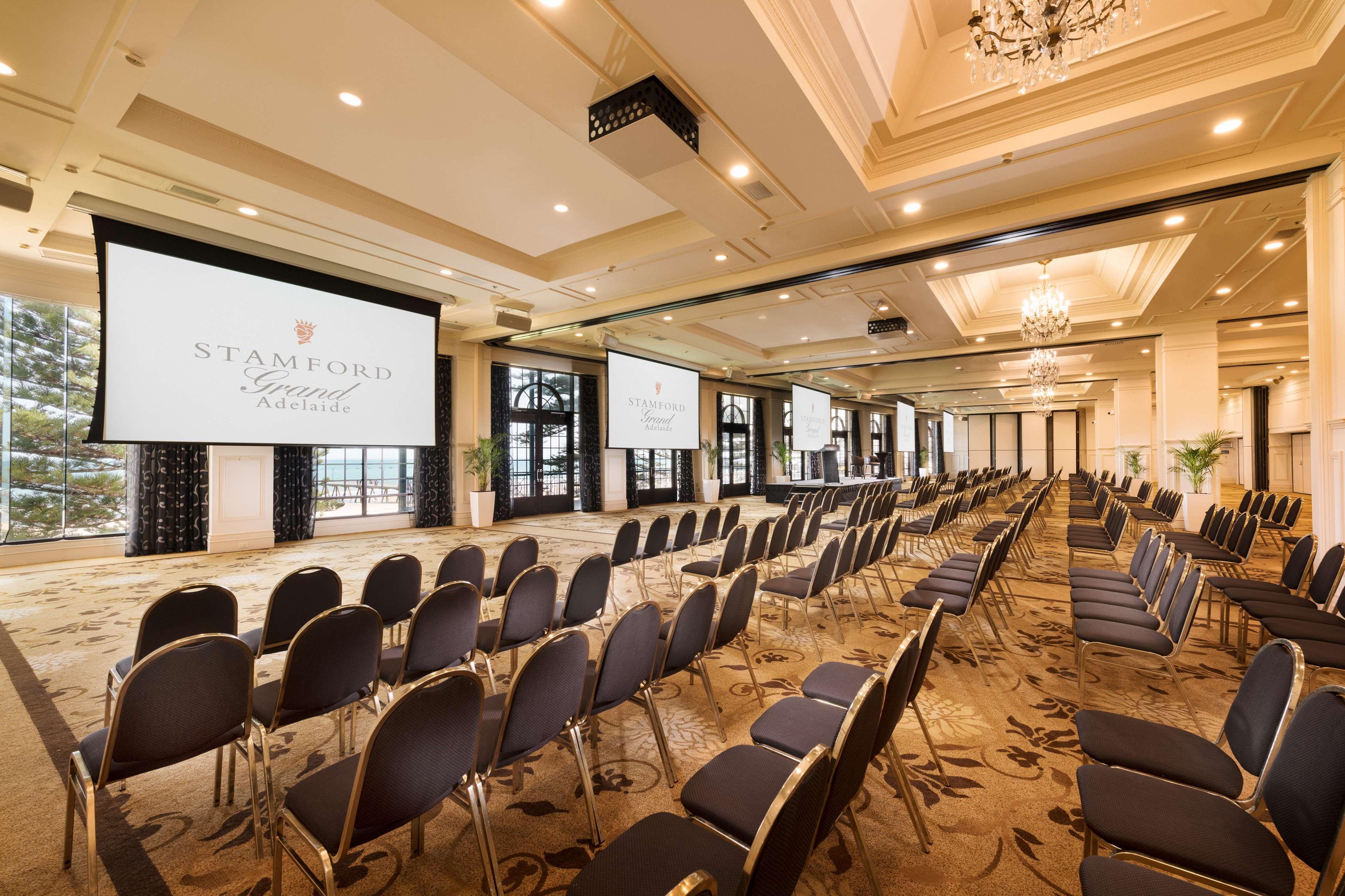 Stamford Grand Adelaide Otel Glenelg Dış mekan fotoğraf The photo shows a spacious event or conference room. There are rows of dark, upholstered chairs arranged facing two large screens. The room features elegant decor with soft lighting, a chandelier, and windows that allow natural light to filter in. Th