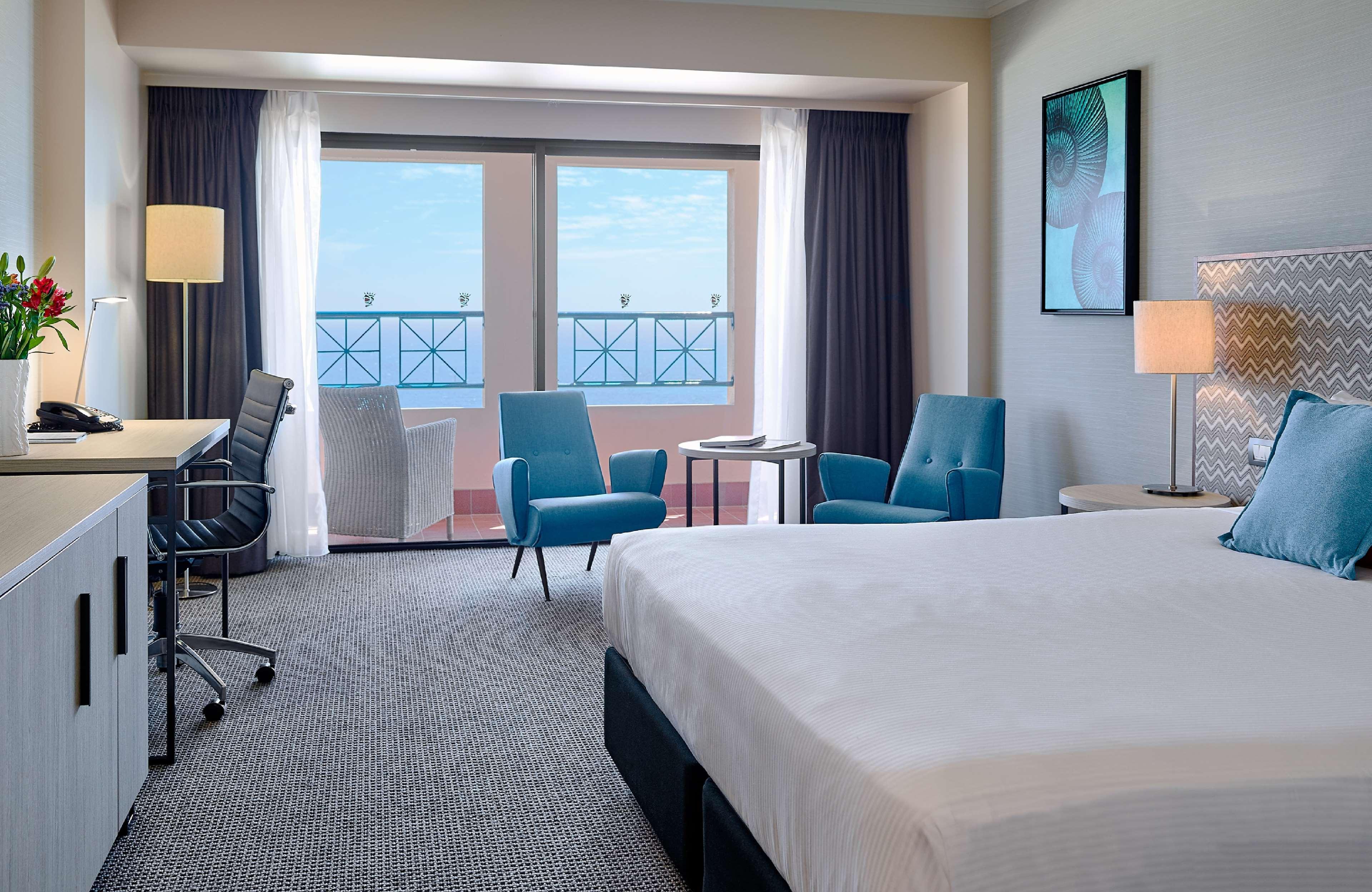 Stamford Grand Adelaide Otel Glenelg Dış mekan fotoğraf The image shows a hotel room featuring a large bed with a white duvet. There are two bright blue chairs and a small table near a large window that provides a view outside, possibly to a balcony. The room includes a workspace with a desk and an office