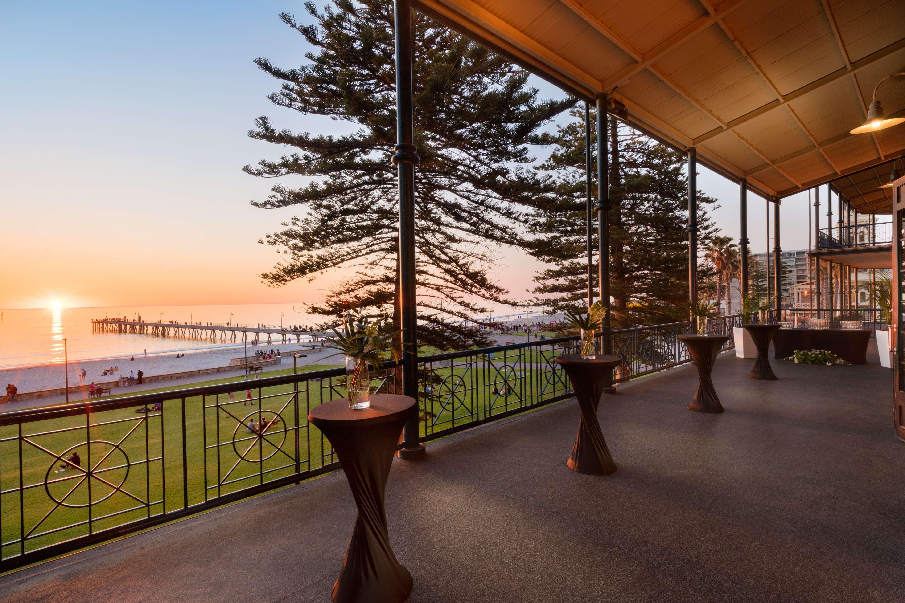 Stamford Grand Adelaide Otel Glenelg Dış mekan fotoğraf The photo shows a spacious indoor space with large windows overlooking a beach at sunset. The scene features a pier extending into the ocean and a horizon painted in warm colors from the setting sun. In the foreground, there are elegant tables with d