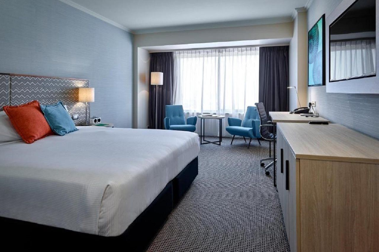 Stamford Grand Adelaide Otel Glenelg Dış mekan fotoğraf The photo shows a modern hotel room with a clean and minimalist design. In the center, there is a large, neatly made bed with white linens and two decorative pillows, one orange and one blue. To the right, there is a small wooden cabinet with a telev